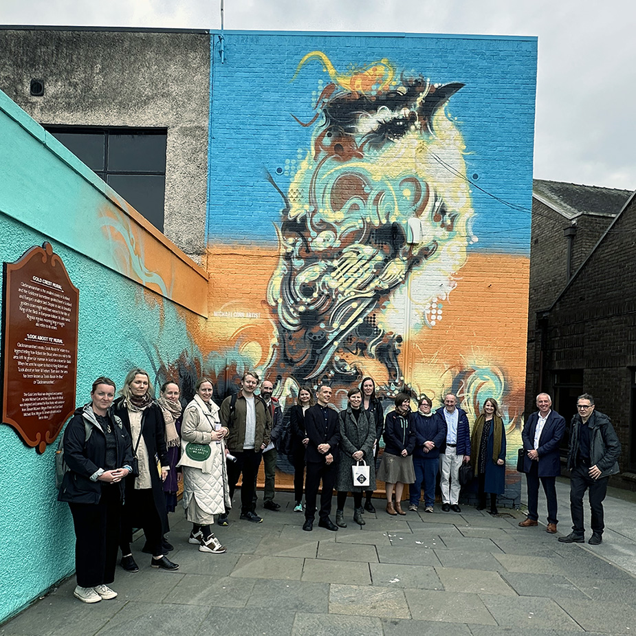 A group of people gather in front of a large colourful mural of a bird.