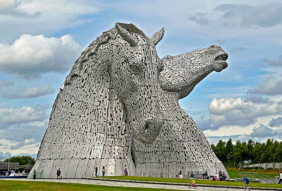 The Kelpies on a sunny day with people cycling and walking around the greenspace around it.