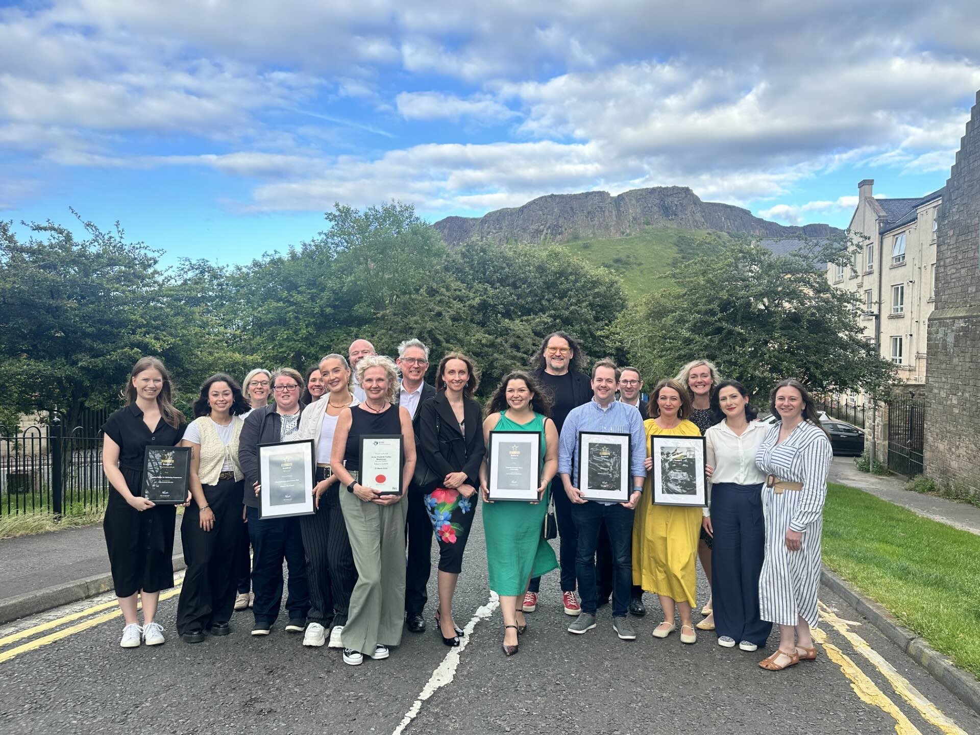A group of people standing in front of Arthurs Seat in Edinburgh holding framed award certificates