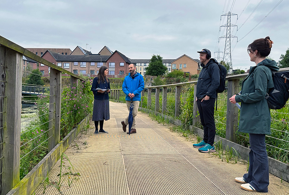 Four people are recording a podcast on a wooden walkway over a canal. The host and the guest are in conversation, while the other two people watch.