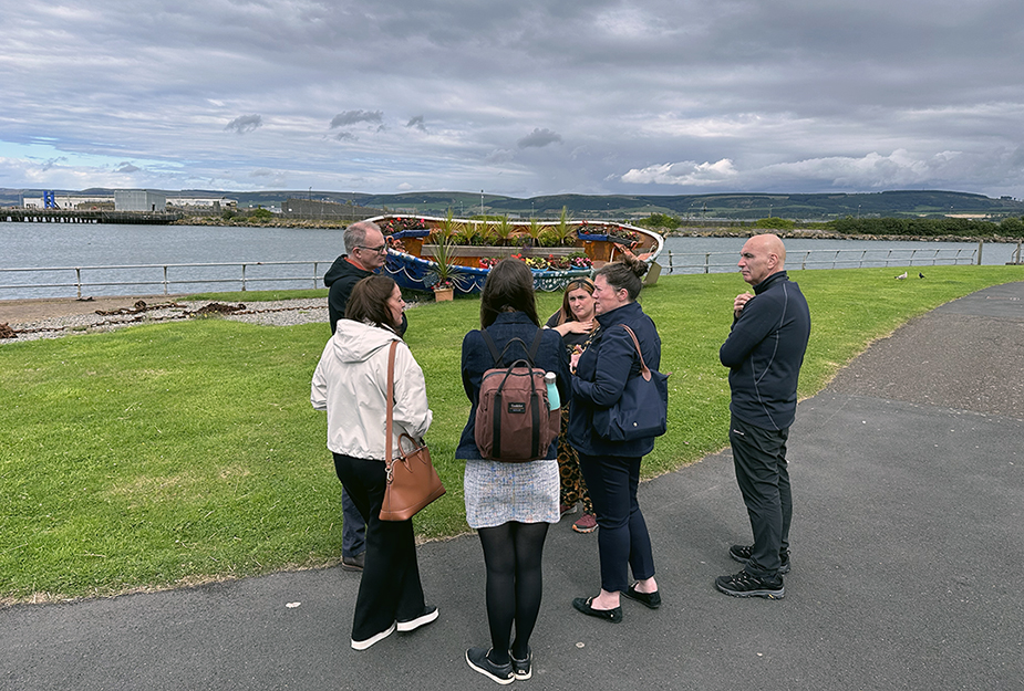 A group of people standing on a sidewalk looking at a garden on a boat.