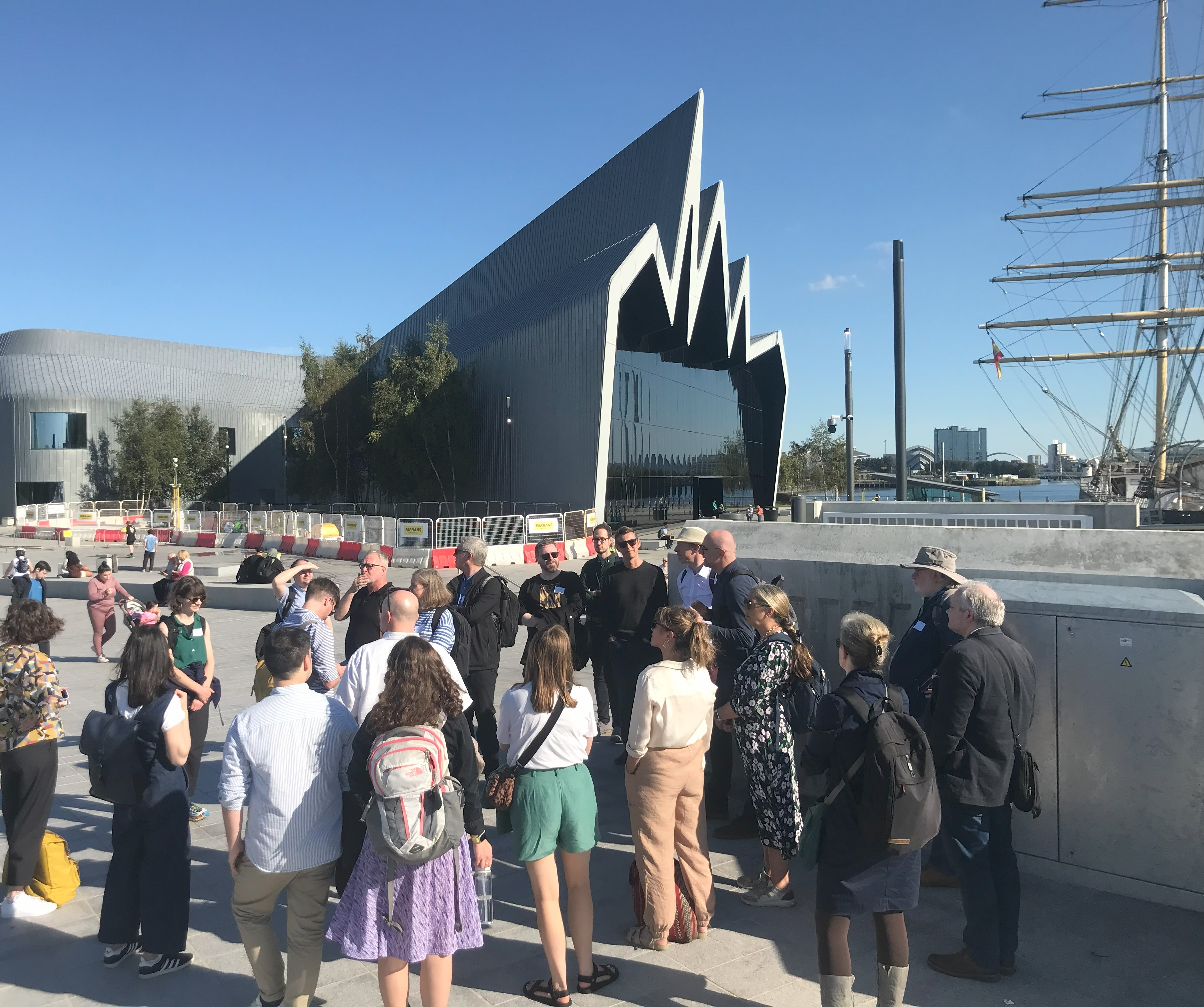 A group of people listen stand outside the Riverside Museum in Glasgow, listening to a tour guide.