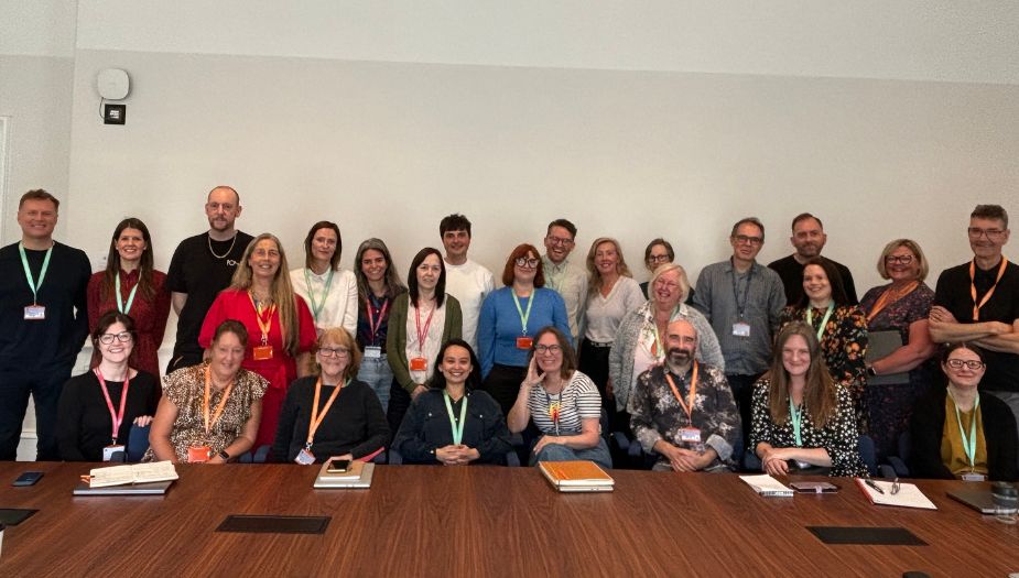 All Architecture and Design Scotland staff pose for a photo together behind a table in a boardroom. 